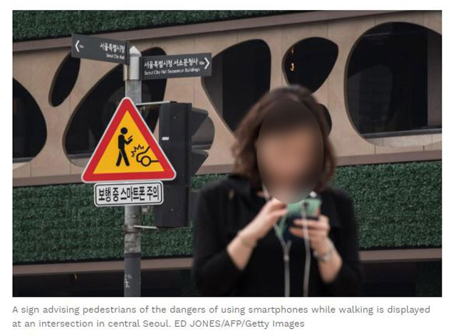 ‘보행 중 스마트폰 주의’라는 표지를 뒤로하고 보행 중 스마트폰을 보고 있는 서울 시민의 모습(출처: Forbes.com)