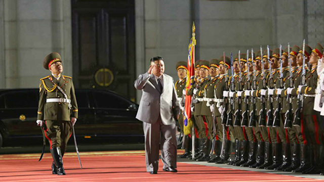 회색 양복을 입은 김정은 위원장이 총을 든 사열대 앞을 지나며 오른손을 들어 경례하고 있다.